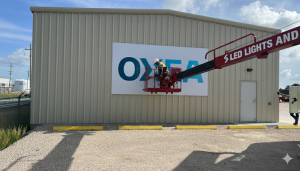 Workers performing Houston sign installation of "Oxygen and LED Lights" on warehouse with boom lift under clear sky.