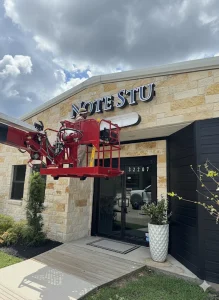 Worker in red scissor lift performing Houston sign installation for "NOTE STU" on stone building under cloudy sky.
