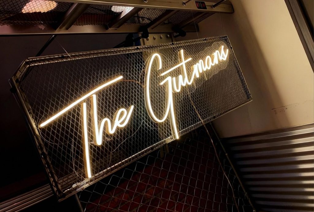 Custom warm-white neon "The Gutmans" building signs Houston with industrial mesh backdrop