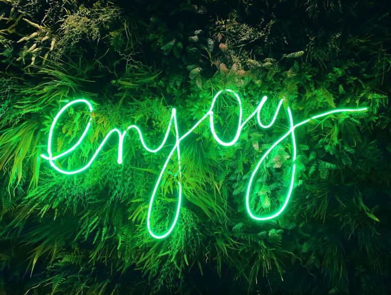 Neon enjoy sign on a greenery wall from a business sign manufacturer Houston specializes in custom lighted signs.