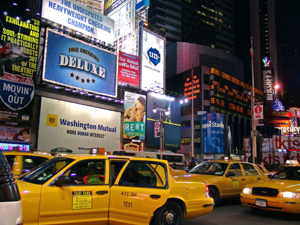 Neon billboards for Houston commercial signs and retail brands light up a busy city street with yellow taxis.